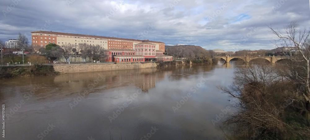 Obraz premium Paisaje urbano junto al río con edificio de ladrillo y puente de piedra antiguo bajo cielo nublado en Logroño, España