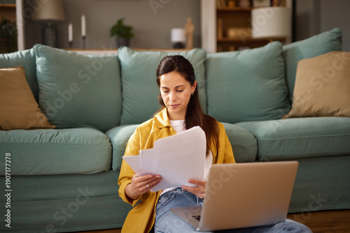Fotografi Woman comfortably sits on the floor in a stylish living room, reading papers while using her laptop on a low table