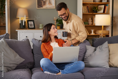 In a cozy living room, a woman smiles at a man while he hands her a cup, both engaged in their relaxed home activities with a laptop.