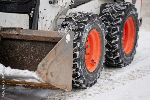 Industrial winter scene of skid steer loader tires with orange rims and deep lugs, with dirty steel bucket in foreground and snow on ground for traction concept