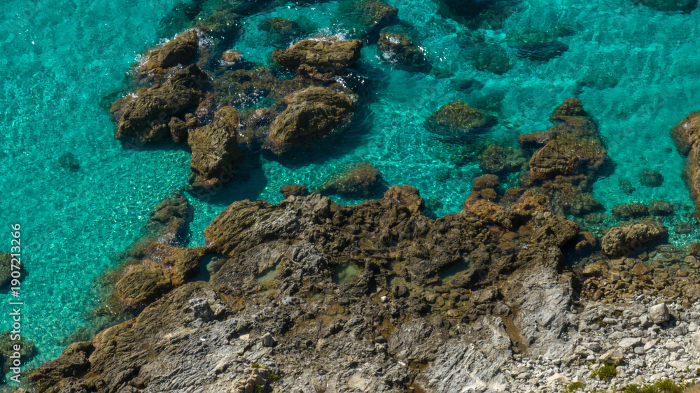 Obraz premium Aerial close-up of crystal-clear turquoise water meeting a rocky shoreline. Large brown rocks are visible beneath the surface, highlighting the incredible transparency of the Mediterranean sea.