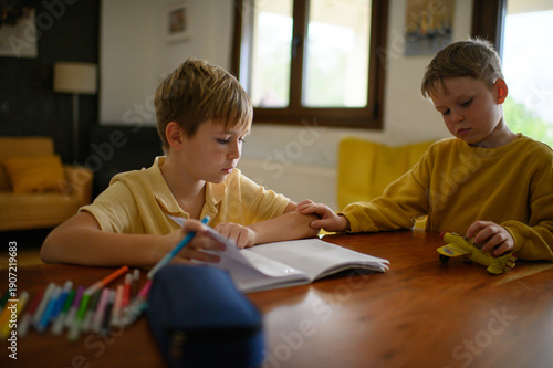 Wallpaper Mural Two boys sitting at a table at home, older brother doing homework and writing, drawing, younger child playing with a toy airplane. Real family lifestyle and everyday childhood scene. Torontodigital.ca
