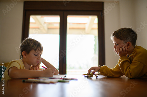 Wallpaper Mural Two brothers sitting at a table at home, one drawing and doing homework while the other plays alone with a toy airplane. Both children look bored in a quiet everyday family scene. Torontodigital.ca