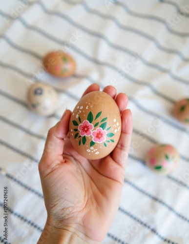 Close-up of a person holding a hand-painted brown Easter egg with delicate pink floral designs, with blurred decorated eggs on a striped linen cloth background.