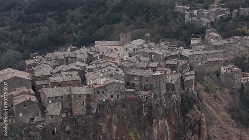 Aerial view of Calcata Vecchia, an ancient medieval village perched on a rocky cliff in Lazio, during a gloomy day. Historic stone houses surrounded by forest and dramatic landscape of a scenic view.
