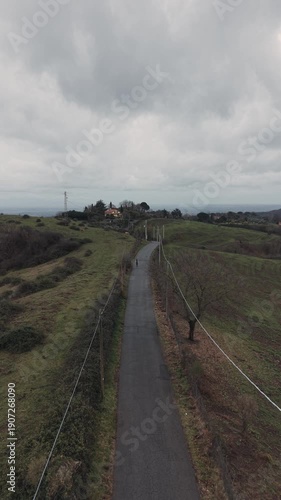 Aerial view of a lone cyclist riding along a narrow countryside road surrounded by rolling green hills under a cloudy sky. Peaceful rural landscape, slow travel concept and outdoor activity.