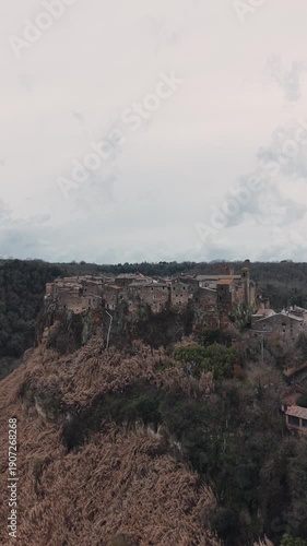 Aerial view of Calcata Vecchia, an ancient medieval village perched on a rocky cliff in Lazio, during a gloomy day. Historic stone houses surrounded by forest and dramatic landscape of a scenic view.
