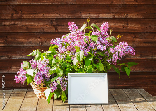 White frame and branches of lilac in vase and in wicker basket on aged rustic wooden table in spring sunny day. View with copy space.