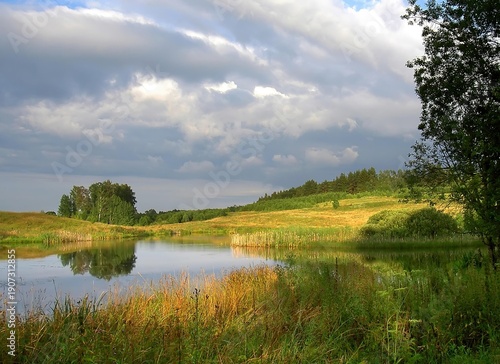 Serene summer landscape featuring a calm lake reflecting lush green hills and a dramatic sky. This tranquil rural scene represents the natural beauty of the Polish countryside.