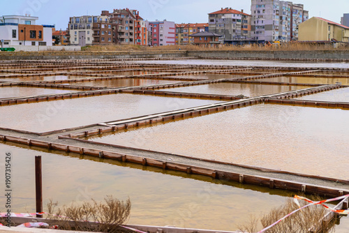 Fotografie Salt evaporation ponds with shallow water, geometric pools near urban area in Pomorie Bulgaria, industrial saltworks landscape