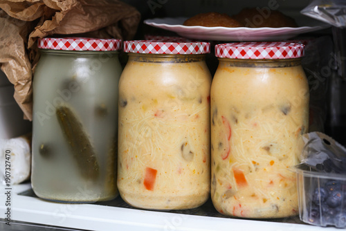 Homemade soups and pickled cucumbers in glass jars inside fridge. Full refrigerator shelf with food containers. Preserved vegetables and soup jars stored together. Cluttered fridge interior closeup.