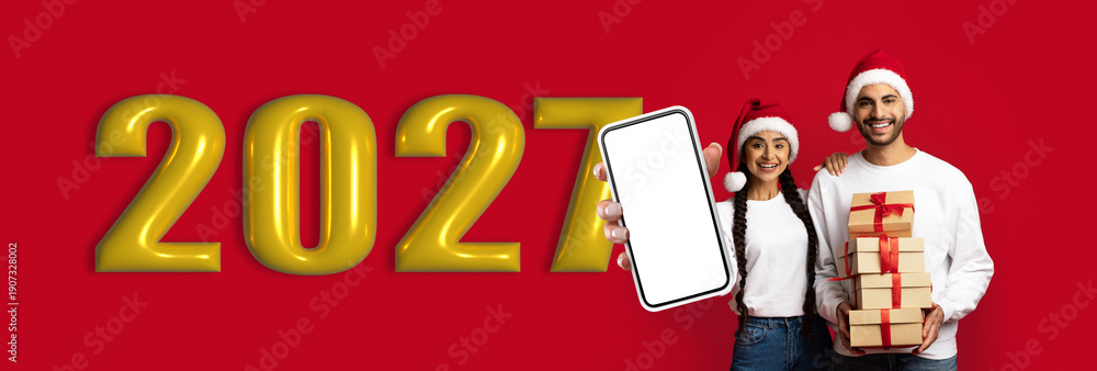 Naklejka premium A couple stands together smiling, wearing Santa hats, with a stack of gift boxes. They are holding a smartphone next to large numbers representing the year 2021 on a red background.