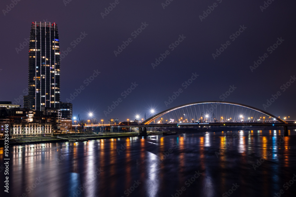 Obraz premium Panorama of Bratislava with illuminated modern buildings on the banks of the Danube and a bridge over the river - against the background of a dark evening sky