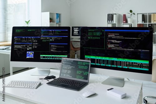Three computer monitors and laptop displaying programming code and system information on desk in office, showing technology workspace with multiple screens and coding activity
