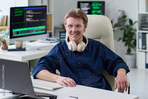 Portrait of young man with glasses and headphones sitting at desk smiling, working in office with computer monitors displaying programming code