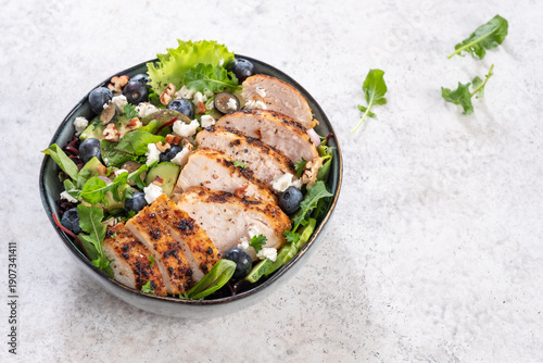 Grilled chicken and blueberry salad with greens and feta cheese arranged in a black bowl on light gray surface