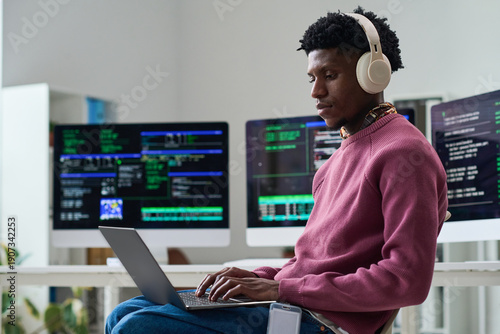 Young Black man wearing headphones working on laptop in front of multiple computer monitors displaying code in office setting, focusing on programming tasks