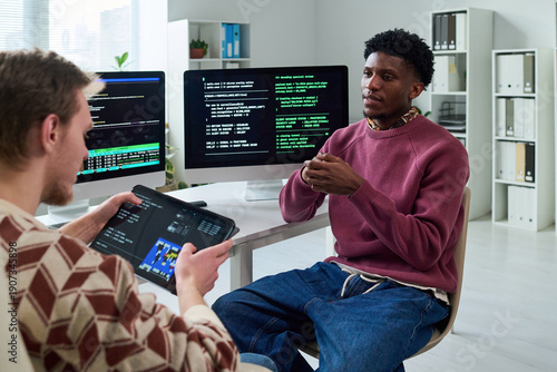 Black young man explaining programming concepts to young man using digital tablet, computer monitors displaying code in office workspace