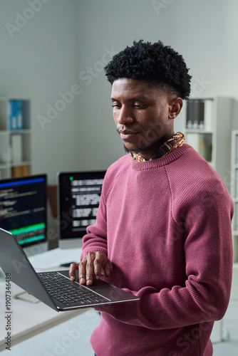 Young Black man standing in office using laptop, looking at computer screen with focused expression, programming or working on software development project