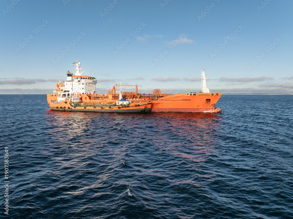Fototapeta premium Aerial drone view of a bright orange industrial chemical tanker ship performing bunkering operations in the blue waters of Baltic Sea.
