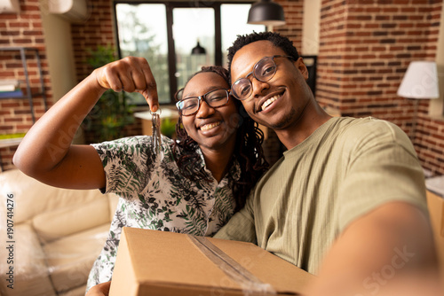 Joyful black couple smiling while holding house keys and cardboard box, excited to move into newly purchased residential property. Black man takes selfie with wife, celebrating fresh start together.