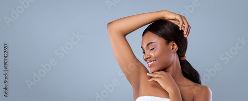 A woman stands against a plain background with her hand raised near her face. She smiles softly with her eyes closed, giving a sense of joy. The setting is minimal and focuses on her expression.