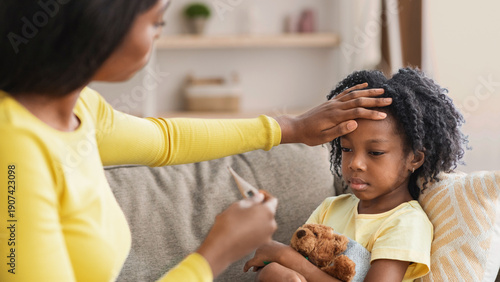 A mother checks the temperature of her sick child who is holding a teddy bear while sitting on a couch. The room is bright with soft furnishings around. It is a comforting moment.