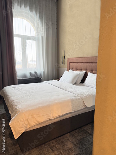 One large double brown bed with white bedding and pillows in a room with a window behind a curtain in the bedroom