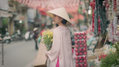 In Hanoi, Vietnam, a woman in traditional dress and a conical hat holds yellow flowers on a busy street. The image captures the essence of Vietnamese culture.
