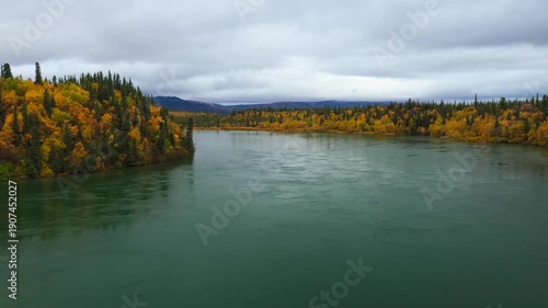 Alaskan wilderness with a flowing river bordered by colorful autumn trees under a cloudy sky