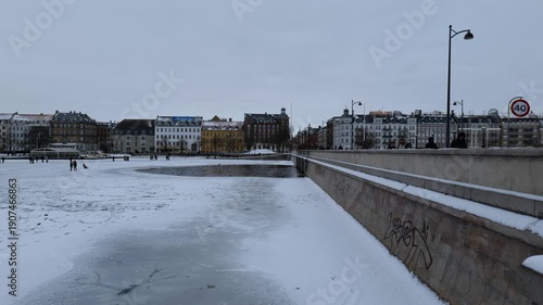 A view from a bridge onto a frozen canal in the city. Many people are walking on the ice. It's very cold and there's a lot of snow