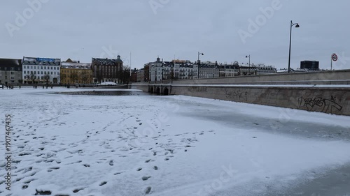 A view from a bridge onto a frozen canal in the city. Many people are walking on the ice. It's very cold and there's a lot of snow