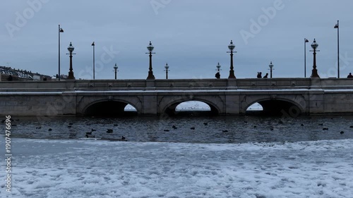 A view across an icy canal to an ancient bridge in the city. Copenhagen in winter. Many ducks swim. People walk