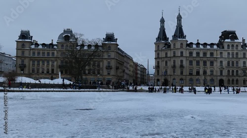 Many people walk on the ice in the middle of a city pond. Beautiful old buildings are in the background. Copenhagen, the capital of Denmark. Cold winter with snow