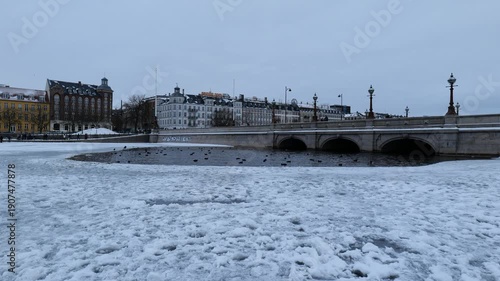 A view across an icy canal to an ancient bridge in the city. Copenhagen in winter. Many ducks swim. People walk