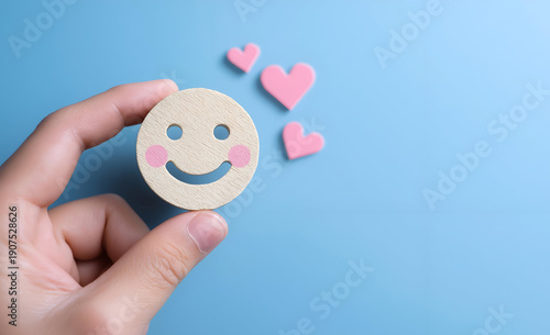 A hand holding a cheerful smiley face cutout with pink hearts, symbolizing joy and positivity against a light blue background.