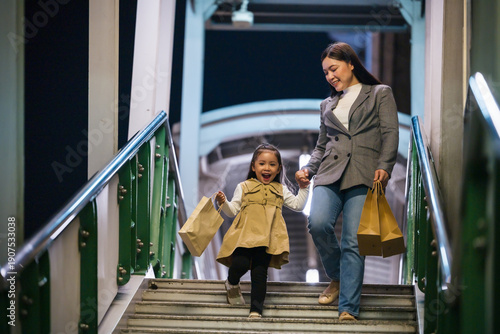 happy mother and child girl with shopping bag enjoying and walking down footbridge stairs in bangkok city at night