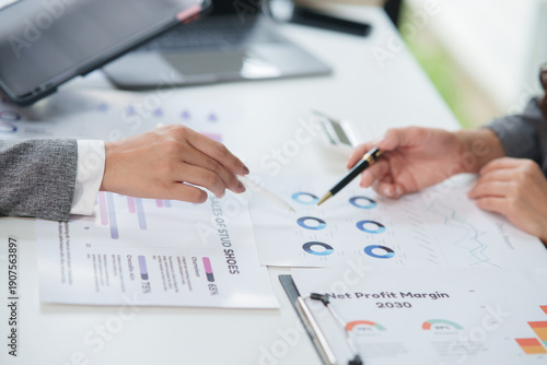Close-up shot of two business professionals' hands pointing and analyzing financial report documents featuring pie charts and bar graphs to evaluate profit margins and business trends in an office set