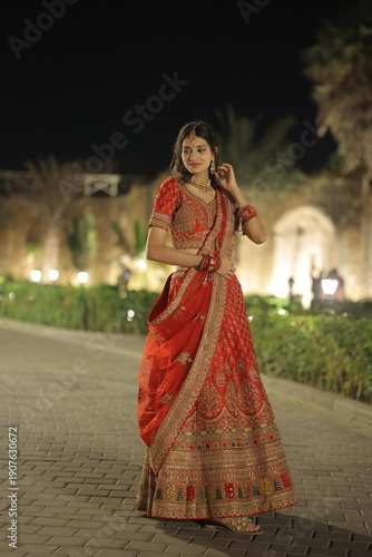Beautiful young Indian woman dressed in an elegant red embroidered lehenga with traditional jewelry, posing outdoors at night