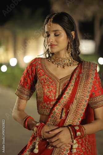 Beautiful young Indian woman dressed in an elegant red embroidered lehenga with traditional jewelry, posing outdoors at night