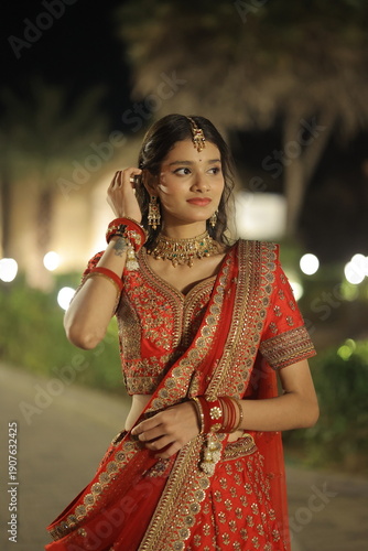 Beautiful young Indian woman dressed in an elegant red embroidered lehenga with traditional jewelry, posing outdoors at night