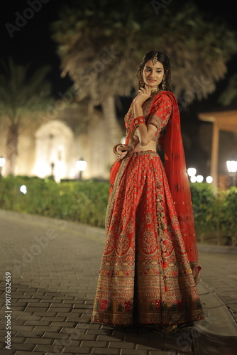Beautiful young Indian woman dressed in an elegant red embroidered lehenga with traditional jewelry, posing outdoors at night