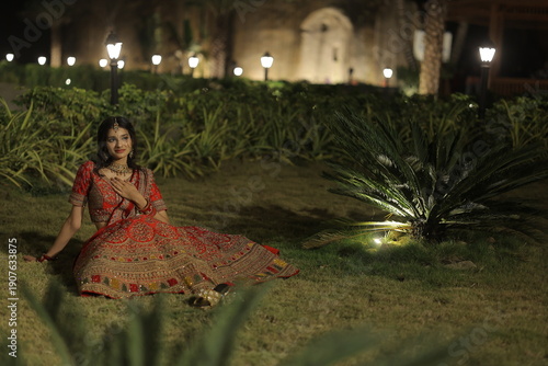 Beautiful young Indian woman dressed in an elegant red embroidered lehenga with traditional jewelry, posing outdoors at night