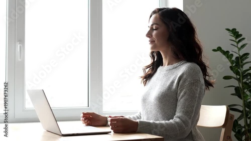 Young woman meditating or relaxing at a desk with a laptop by a bright window in a home office setting.