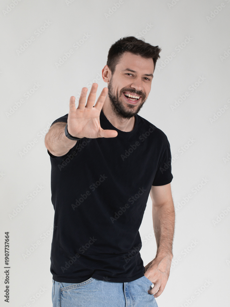 Fototapeta premium Studio shot of a cheerful millennial man with a beard, in a black t-shirt and blue jeans. He laughs heartily while extending one hand towards the camera in a stop or high-five gesture.