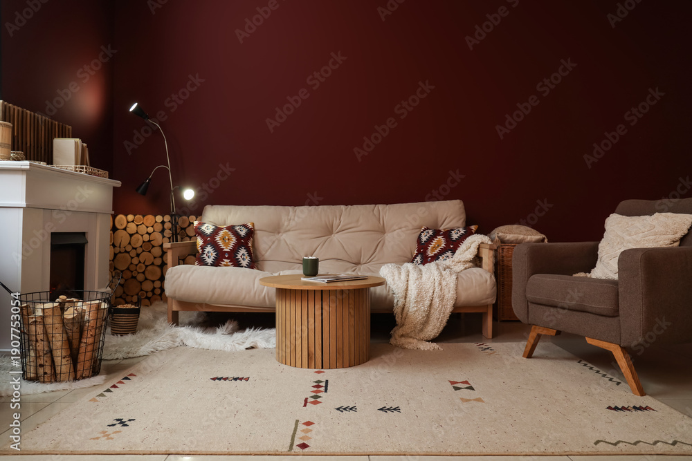 Naklejka premium Interior of living room with sofa, armchair, coffee table, fireplace and firewood in basket near burgundy wall