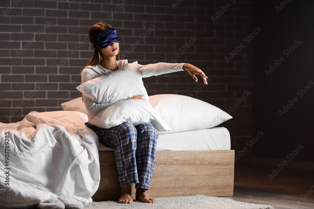 Fototapeta premium Female sleepwalker with sleeping mask and pillow sitting on bed near black brick wall in dark bedroom at night