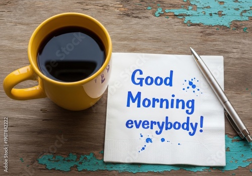 A bright yellow coffee mug sits beside a white napkin with 'Good Morning everybody!' written in blue ink.