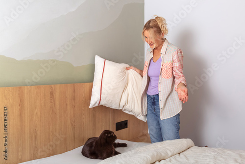 A woman with a smile looks at a cat that has lain down on the bed and does not let her make the bed.