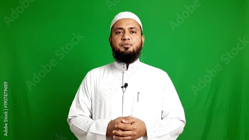 A Muslim man wearing a white thobe and cap standing in front of a green screen during ramadan with a microphone.